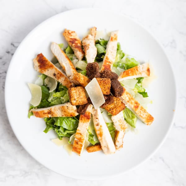 Overhead view of a Caesar salad with grilled chicken, croutons, and parmesan cheese on a white plate.