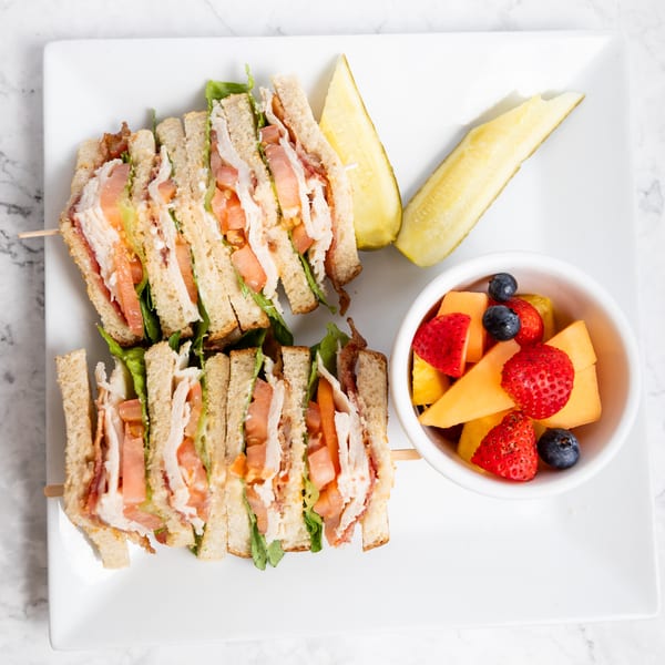 Club sandwich with turkey, bacon, lettuce, and tomato, served with pickles and fruit salad.