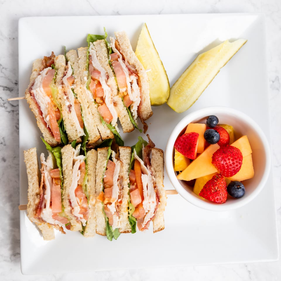 Club sandwich with turkey, bacon, lettuce, and tomato, served with pickles and fruit salad.