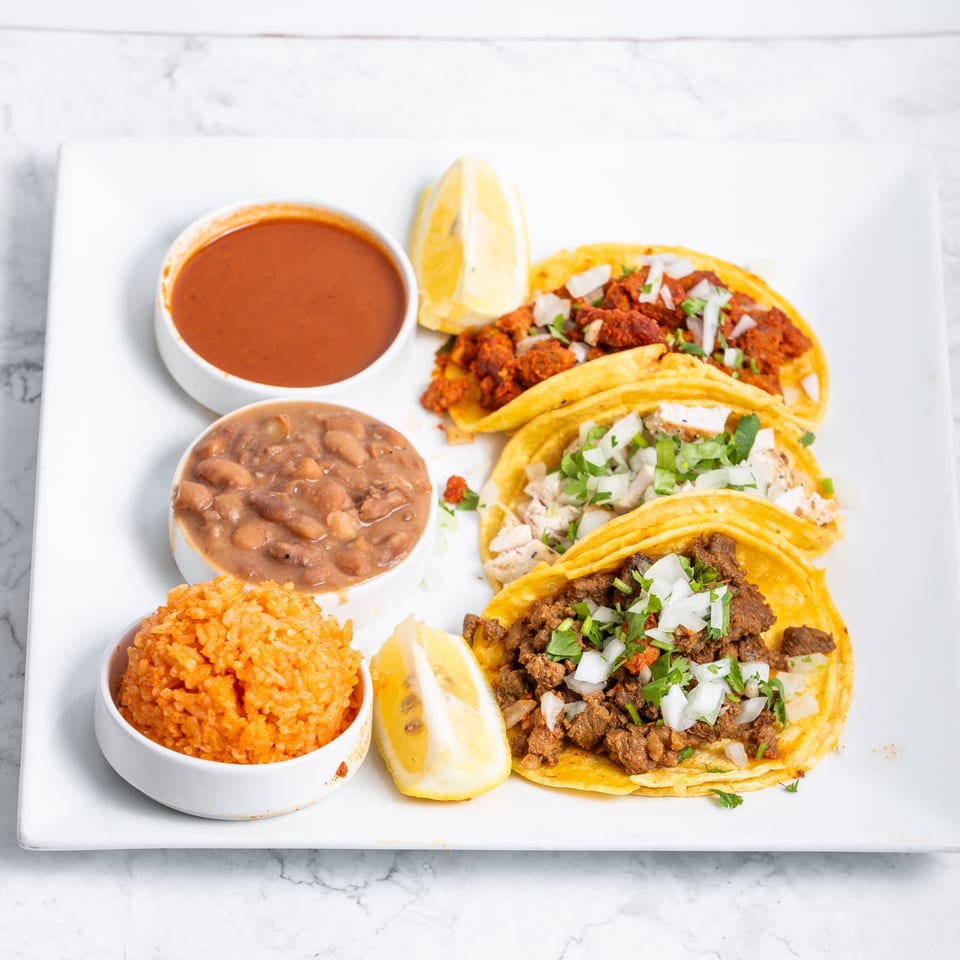 Three tacos with various fillings, rice, beans, and salsa, garnished with lemon wedges.