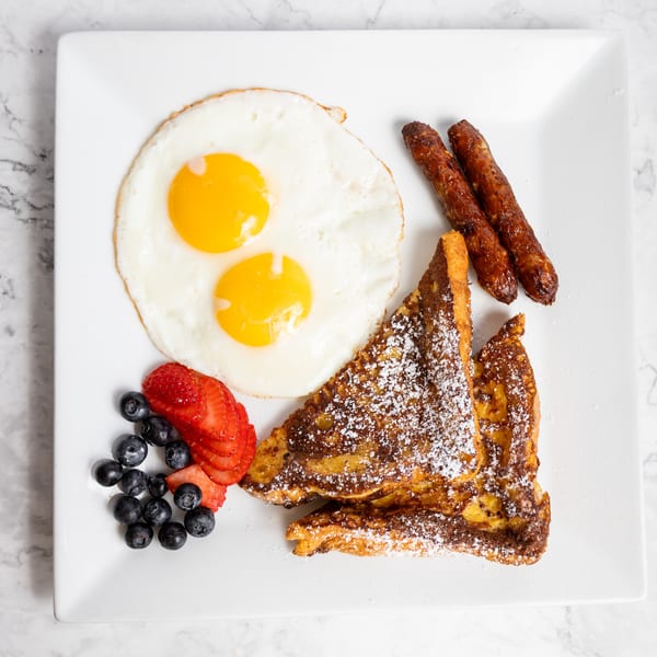 Breakfast plate with two fried eggs, two sausage links, French toast, blueberries, and sliced strawberries.